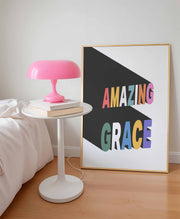 Poster featuring the text 'AMAZING GRACE' in colorful letters on a white background, framed and displayed beside a pink lamp.