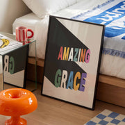 Framed poster with colorful text 'AMAZING GRACE' on a white background, placed on a side table next to a red mug.