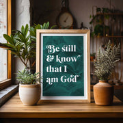 Botanical poster with the text 'Be still and know that I am God' surrounded by green leaves, framed on a wooden table.