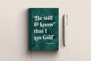 A5 hardback journal with green leaf design and white text reading 'Be still & know that I am God' Psalm 46:10, next to a pen.