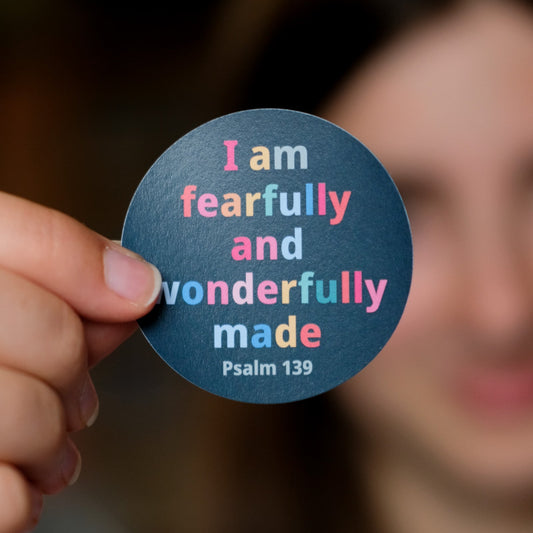 A close-up of a hand holding a round sticker with the text 'I am fearfully and wonderfully made Psalm 139' in pastel pink, blue, and yellow lettering on a dark blue background. The background is heavily blurred, showing a person's face and hair out of focus.