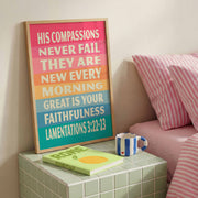 Colorful poster with text from Lamentations 3:22-23 on a bedside table next to a striped pillow and a cup.