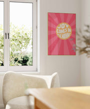 Poster with pink background and white text reading 'Joy Comes in the Morning', featuring sunburst design.