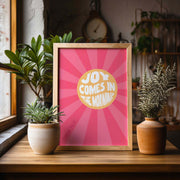 Poster with the text 'JOY COMES IN THE MORNING' in a circular design, set against a pink sunburst background, framed.