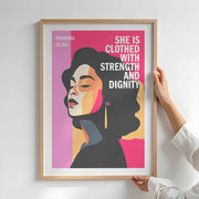 Framed poster featuring a woman with text: 'She is clothed with strength and dignity' on a pink background.