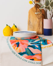 Organic cotton tea towel with floral design and Matthew 6 quote, styled with plates and decorative items.
