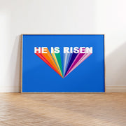 Colorful poster with the text 'HE IS RISEN' on a blue background, framed and displayed on a wooden floor.