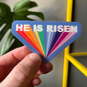 Eco-vinyl sticker featuring the text 'HE IS RISEN' in colorful, bold letters on a blue background.