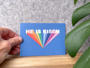 Colorful Easter postcard with the text 'HE IS RISEN' on a blue background, held near a green plant.