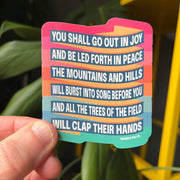 Colorful eco-vinyl sticker featuring the text of Isaiah 55:12 with a joyful design, held against a plant background.