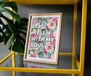 Framed poster with floral design and text 'IT IS WELL WITH MY SOUL' on a yellow shelf with a plant.