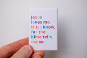 A hand holding a magnet with colorful text: 'Jesus loves me, this I know, for the bible tells me so.