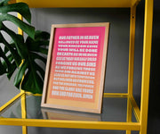 Framed poster featuring the Lord's Prayer in bold pink and orange text, displayed on a yellow shelf with greenery.