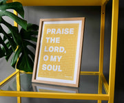 Framed poster with yellow background displaying the text 'PRAISE THE LORD; O MY SOUL' from Psalm 103, set on a yellow shelf.