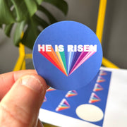Circular sticker with the text 'HE IS RISEN' in colorful letters on a blue background, held in a hand.