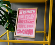 Framed poster with pink text: 'Strength for today and bright hope for tomorrow' on a yellow shelf with a plant.