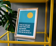 A framed poster with a teal background, yellow sun, and text: 'This is the day that the Lord has made. Let us rejoice and be