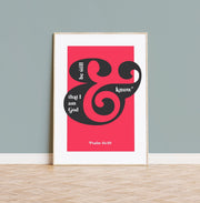 Framed poster with bold black text on a pink background, featuring the phrase 'be still and know that I am God' and 'Psalm