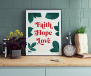 Poster featuring the words 'Faith Hope Love' in bold red letters surrounded by green leaves, displayed on a wooden table.