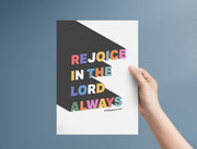 Hand holding a poster with colorful text: 'Rejoice in the Lord Always' and Philippians 4:4, against a blue background.