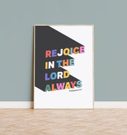 Colorful poster featuring the text 'REJOICE IN THE LORD ALWAYS' with Philippians 4:4, framed against a light wall.