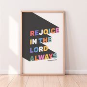 Colorful poster with the text 'Rejoice in the Lord Always' and Philippians 4:4, framed against a light wall.