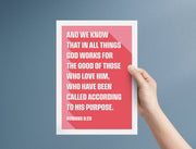 Poster featuring Romans 8:28 in bold white text on a pink background, held against a blue wall.