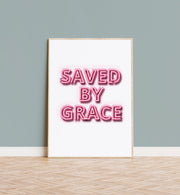 Neon effect poster with the text 'SAVED BY GRACE' in pink on a white background, framed and displayed on a wall.