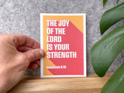 Postcard featuring the text 'The Joy of the Lord is your strength' with Nehemiah 8:10, held against a gray background.